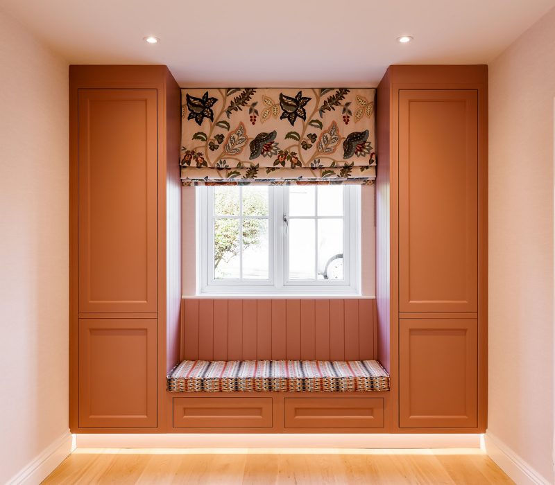 Bespoke Kitchens Near me Window seat with a striped cushion between two tall terracotta-coloured cupboards